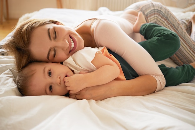 Piękna blondynka uśmiechnięta szeroko leżąca na rozłożonym łóżku i obejmująca przebudzonego malucha. Przytulne słodkie ujęcie uroczej mamy i małego dziecka w sypialni. Rodzina, miłość, troska i uczucie