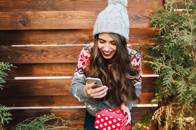 Piękna brunetka dziewczyna z długimi włosami i czerwonymi ustami na drewnianym zewnątrz. Nosi dzianinową czapkę, trzymając telefon i pudełko z prezentami. Wygląda na szczęśliwą.