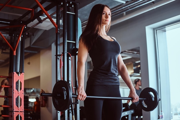 Piękna brunetka kobieta w odzieży sportowej trzyma sztangę podczas treningu w klubie fitness lub siłowni.