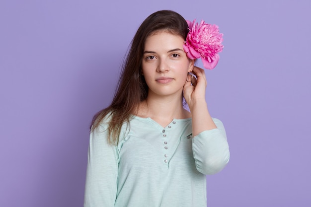Piękna brunetki młoda kobieta jest ubranym przypadkową odzież trzyma różowego peonia kwiatu za jej ucho