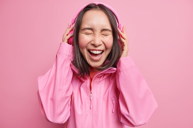 Piękna ciemnowłosa Azjatka słucha zabawnej historii online przez słuchawki stereo, śmieje się radośnie zamyka oczy z radości, nosi kurtkę odizolowaną na różowej ścianie. Koncepcja stylu życia ludzi emocje
