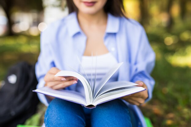 Piękna ciemnowłosa poważna dziewczyna w dżinsowej kurtce i okularach czyta książkę w parku.