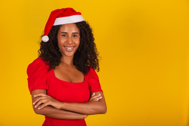Piękna czarna kobieta afro z kręconymi włosami na żółtym tle w santa hat z rękami skrzyżowanymi i wolnym miejscem na tekst uśmiechający się patrząc na kamery