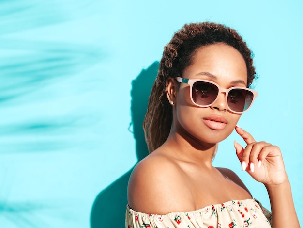 Piękna czarna kobieta z fryzurą afro loki Uśmiechnięta modelka ubrana w letnie ubrania hipster Seksowna beztroska kobieta pozuje przy niebieskiej ścianie w studio Opalona i wesoła w okularach przeciwsłonecznych
