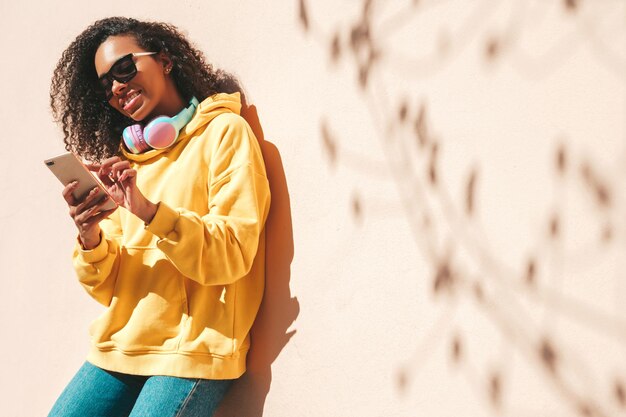 Piękna czarna kobieta z fryzurą afro loki Uśmiechnięta modelka w żółtej bluzie z kapturem Seksowna beztroska kobieta pozuje na tle ulicy w okularach przeciwsłonecznych Patrząc na ekran smartfona za pomocą aplikacji