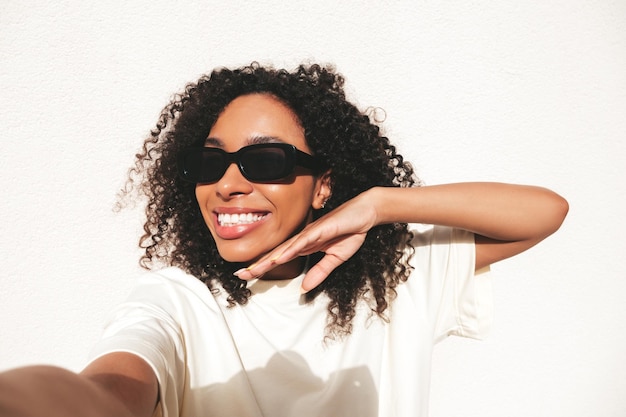 Piękna czarna kobieta z fryzurą afro lokiUśmiechnięta modelka hipster w białej koszulce Seksowna beztroska kobieta pozuje na ulicy w pobliżu białej ściany w okularach przeciwsłonecznych Wesoła i szczęśliwaTworzenie selfie POV