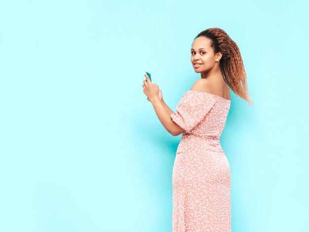 Piękna czarna kobieta z fryzurą afro lokiUśmiechnięta modelka w modnych letnich ubraniach Seksowna beztroska kobieta pozuje w pobliżu niebieskiej ściany w studioKorzystanie z aplikacji na smartfoniePatrząc na ekran telefonu komórkowego