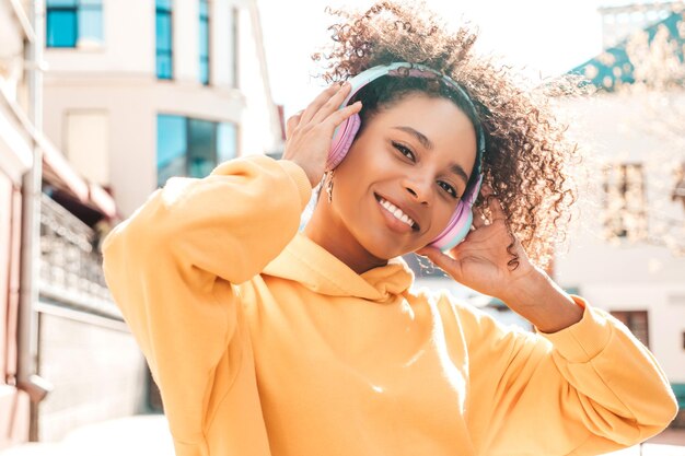 Piękna czarna kobieta z fryzurą afro lokiUśmiechnięta modelka w żółtej bluzie z kapturemSeksowna beztroska kobieta słuchająca muzyki w bezprzewodowych słuchawkachPozowanie na tle ulicy o zachodzie słońca