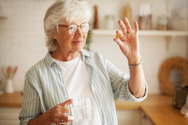 Piękna dojrzała sześćdziesięcioletnia kobieta w stylowych okularach trzymająca kubek i kapsułkę z suplementem omega 3, zamierzająca zażywać witaminę po posiłku. Starszy siwy kobieta bierze pigułkę oleju z ryb wodą