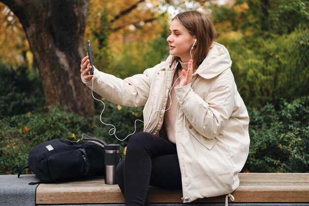 Piękna dziewczyna w puchowej kurtce, machając gestem „Hi”, rozmawiając na czacie wideo na telefonie komórkowym w parku miejskim