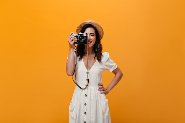 Piękna dziewczyna w stylowym letnim stroju robi zdjęcie aparatem retro. Modna kobieta w białej długiej sukni i kapeluszu z uśmiechem.