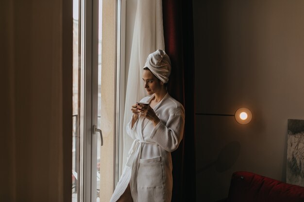 Piękna dziewczyna w szlafroku i ręczniku na głowie wygląda w zamyśleniu przez okno z filiżanką herbaty w dłoniach.