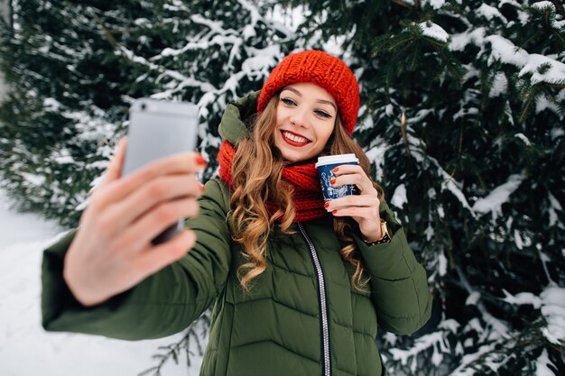 Piękna dziewczyna w zimowe ubrania ma selfie na smartphone. Kobieta z kawą bierze