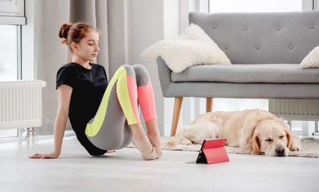 Piękna dziewczyna wyciąga palce u nóg podczas lekcji baletu online z tabletem w domu i śpiącym obok niej psem golden retriever
