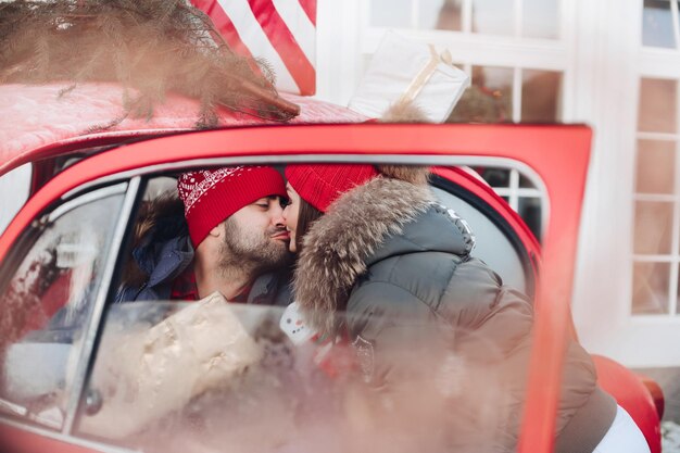 Piękna kaukaska dziewczyna w ciepłych zimowych ubraniach niesie swojemu mężowi pudełka ze świątecznymi prezentami w czerwonym samochodzie