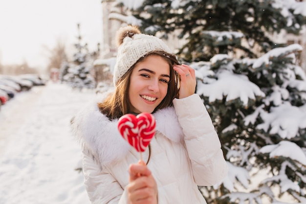 Piękna kaukaski kobieta z czerwonym cukierkiem, ciesząc się zimą na wakacjach. Plenerowe zdjęcie zrelaksowanej kobiety w białej czapce z dzianiny, pozującej na ulicy ze śniegiem