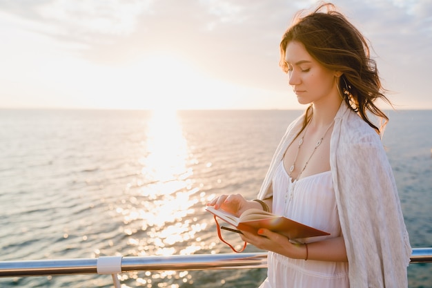 Bezpłatne zdjęcie piękna kobieta w białej letniej sukience spaceru nad morzem o wschodzie słońca z książką do pamiętnika w romantycznym nastroju, myśląc i robiąc notatki
