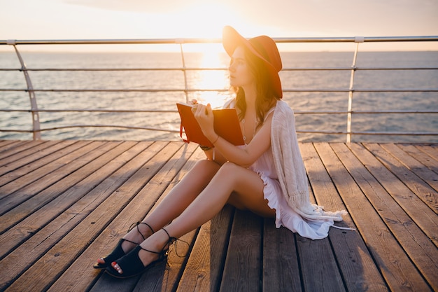 Piękna kobieta w białej sukni siedzi nad morzem na wschód słońca, myśląc i robiąc notatki w dzienniku