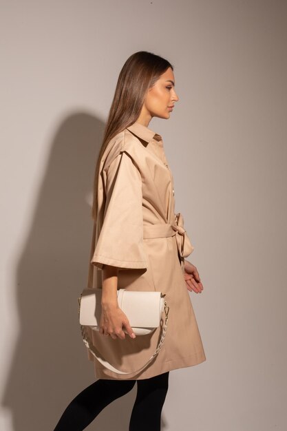 Piękna kobieta w brązowym płaszczu, czarnych spodniach, białych butach, trzyma białą torbę pozującą do aparatu
