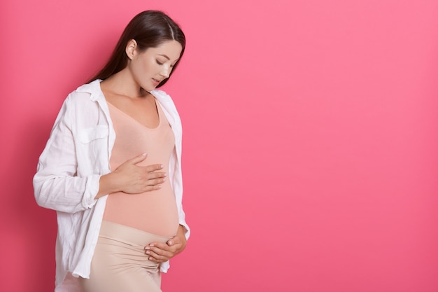 Piękna kobieta w ciąży przytula jej brzuch przed różową przestrzenią, patrząc na jej brzuch z miłością, skopiuj miejsce na reklamę lub tekst promocyjny.