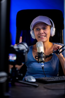 Piękna kobieta w czapce i białych słuchawkach prowadzi transmisję na żywo przez internet. mówi do mikrofonu. blogger na żywo rozmawia z kamerą na komputerze z monitorami