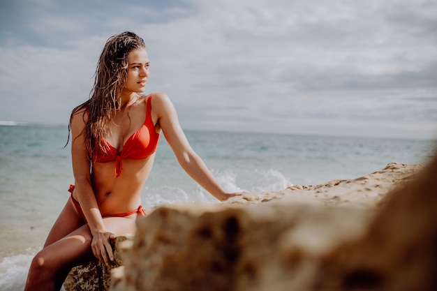 Piękna kobieta w czerwonym bikini pozowanie na plaży, siedząc na skałach