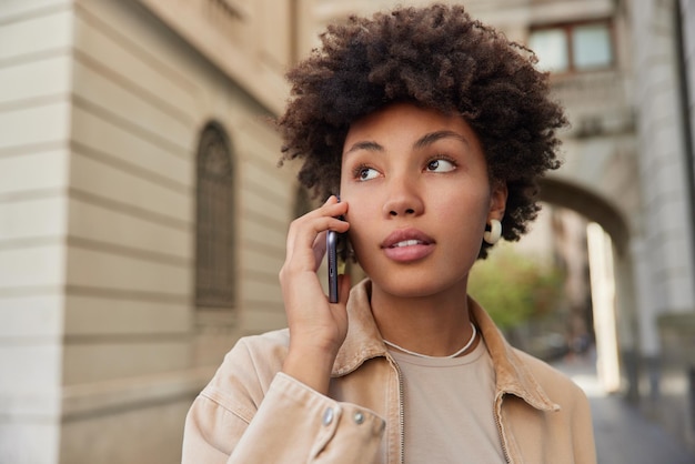 Bezpłatne zdjęcie piękna kobieta z kręconymi włosami sprawia, że dzwoni telefon, spaceruje po mieście, nosi beżową kurtkę, odwraca wzrok z zamyślonym wyrazem twarzy omawia najnowsze wiadomości ludzie koncepcja stylu życia i wypoczynku