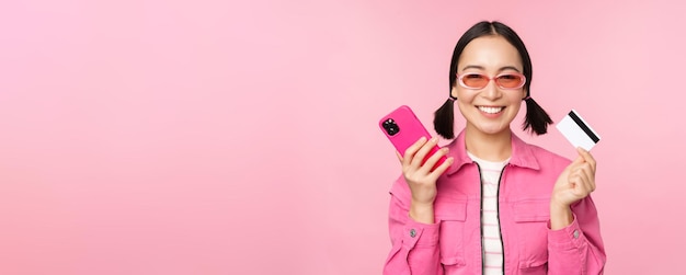 Piękna koreańska kobieta trzymająca kartę kredytową smartfona uśmiecha się do kamery kupując zakupy online z telefonem komórkowym stojącym na różowym tle
