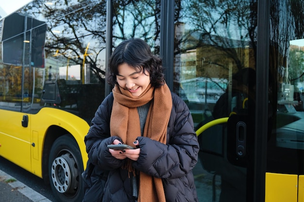 Piękna koreańska studentka na przystanku autobusowym patrząca na swój smartfon sprawdzający tekst do czytania w rozkładzie jazdy