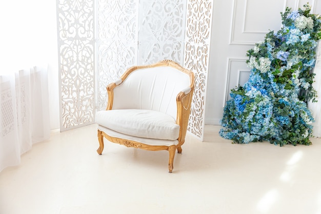 Piękna luksusowa klasyczna biała jasna, czysta sypialnia w stylu barokowym z dużym oknem, fotelem i kompozycją kwiatową.