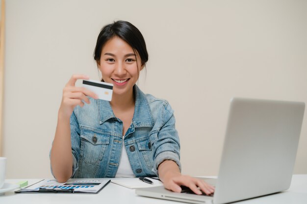 Piękna mądrze biznesowa Azjatycka kobieta używa komputer lub laptop kupuje online zakupy kredytową kartą podczas gdy będący ubranym mądrze przypadkowego obsiadanie na biurku w żywym pokoju w domu.
