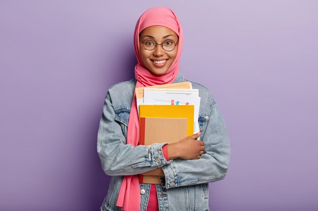 Piękna młoda Arabka o ciemnej karnacji, nosi przezroczyste okulary, trzyma w ręku papiery i notes, ma zębaty uśmiech