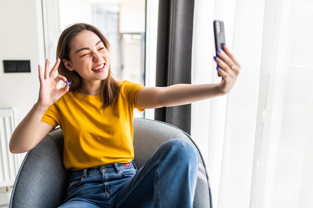 Piękna młoda azjatycka kobieta robi selfie swoim smartfonem i uśmiecha się siedząc w dużym wygodnym fotelu w domu