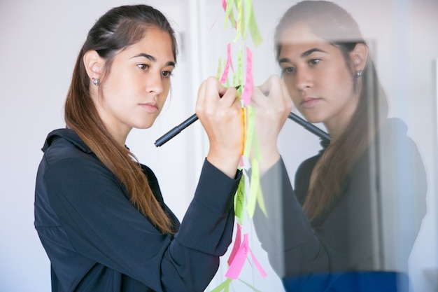 Bezpłatne zdjęcie piękna młoda bizneswoman latin pisze na naklejce z markerem. skoncentrowana profesjonalna menadżerka, dzieląca się pomysłem na projekt i notująca