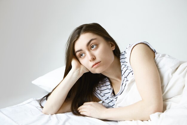 Piękna młoda brunetka Europejka w pasiastej piżamie czuje się samotna i znudzona na śmierć, leżąc w łóżku, patrząc w bok z zamyślonym wyrazem twarzy
