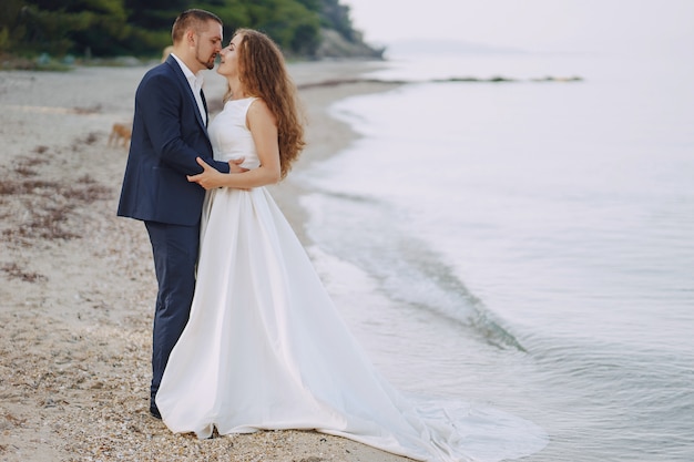 piękna młoda długowłosa panna młoda w białej sukni z mężem na plaży