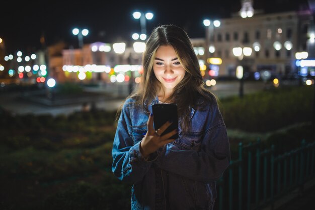 Piękna młoda dziewczyna SMS-y na telefon komórkowy na zewnątrz nad niewyraźną nocną ścianą ulicy, selektywne focus