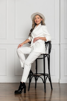 Piękna młoda dziewczyna z piękną sylwetką w białym garniturze spodni i białym kapeluszu z szerokim rondem pozuje na białym tle w studio. siedząc na krześle