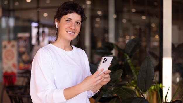 Piękna młoda kaukaski brunetka dziewczyna w białym swetrze patrzy na kamerę, siedząc w sieciach społecznościowych przez telefon w kawiarni