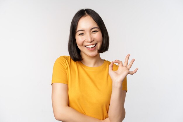 Piękna młoda kobieta pokazująca znak OK, uśmiechając się, zadowolona, polecając coś zatwierdzić, jak produkt stojący w żółtej koszulce na białym tle