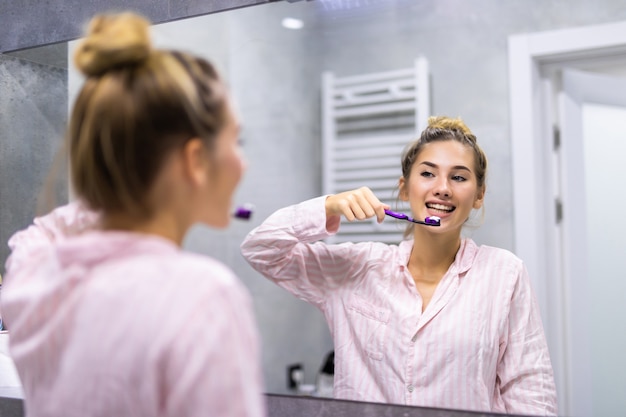 Piękna młoda kobieta szczotkuje zęby przed lustrem w łazience. Selektywne skupienie.