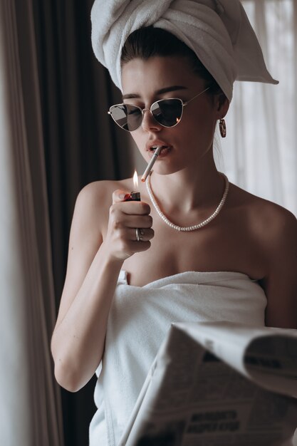 Piękna młoda kobieta w ręczniku pali papierosa i czyta gazetę