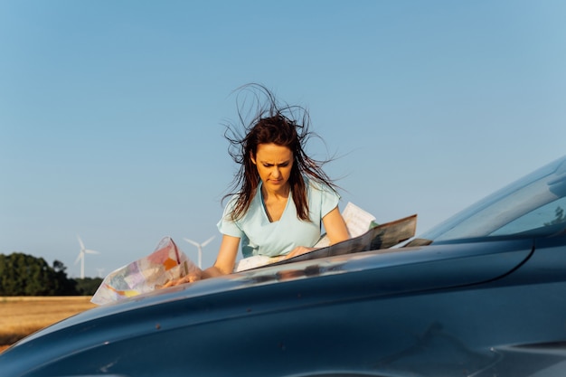Piękna młoda kobieta w średnim wieku patrząc na mapę na szczycie samochodu z silnym wiatrem i wiatrakami w tle. lokalizowanie celu.