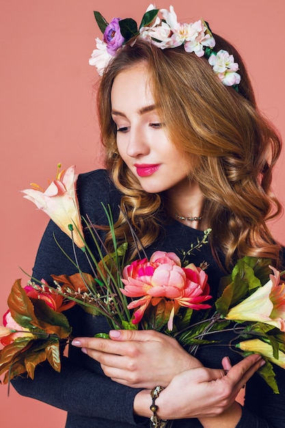 Piękna młoda kobieta z długie falowane blond włosy w wieniec wiosennych kwiatów z bukietem kwiatów na różowym tle.