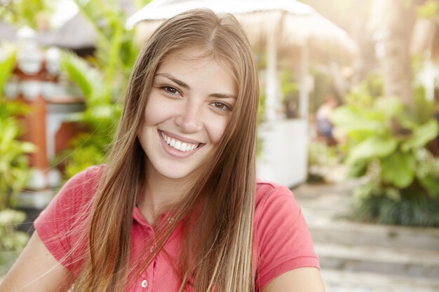 Piękna młoda kobieta z długimi jasnymi włosami ubrana w koszulkę polo, patrząc i uśmiechając się z radosnym wyrazem radości, stojąc na zewnątrz przed zielonymi roślinami