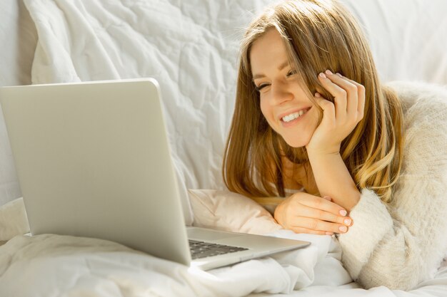 Piękna młoda kobieta za pomocą swojego laptopa, leżąc na kanapie w domu w ciepłym świetle słonecznym.