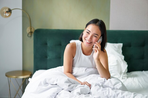 Bezpłatne zdjęcie piękna młoda koreańska kobieta dzwoni, rozmawiając przez telefon w łóżku z przyjacielem podczas pobytu w domu