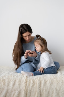Piękna młoda matka i jej córeczka siedzą w domu na łóżku. smartfon telefonu komórkowego w ręce. kobieta pracuje, dręczy ją dziewczyna. miejsce na tekst
