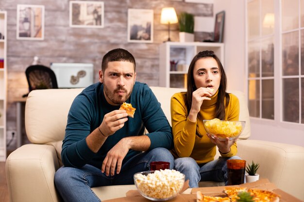 Piękna młoda para ogląda telewizję i je fast food na wynos w salonie