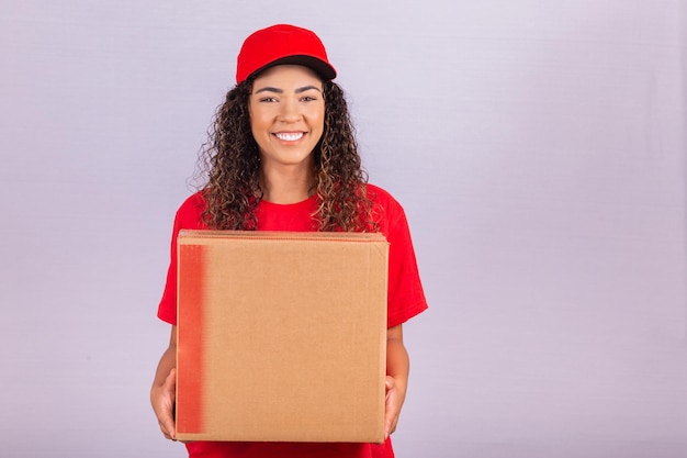 Piękna młoda posłanka dostarczająca ogromną paczkę. doręczycielka w czerwonym mundurze uśmiecha się z dużym kartonowym pudełkiem w dłoniach. zakupy online. równość kobiet.